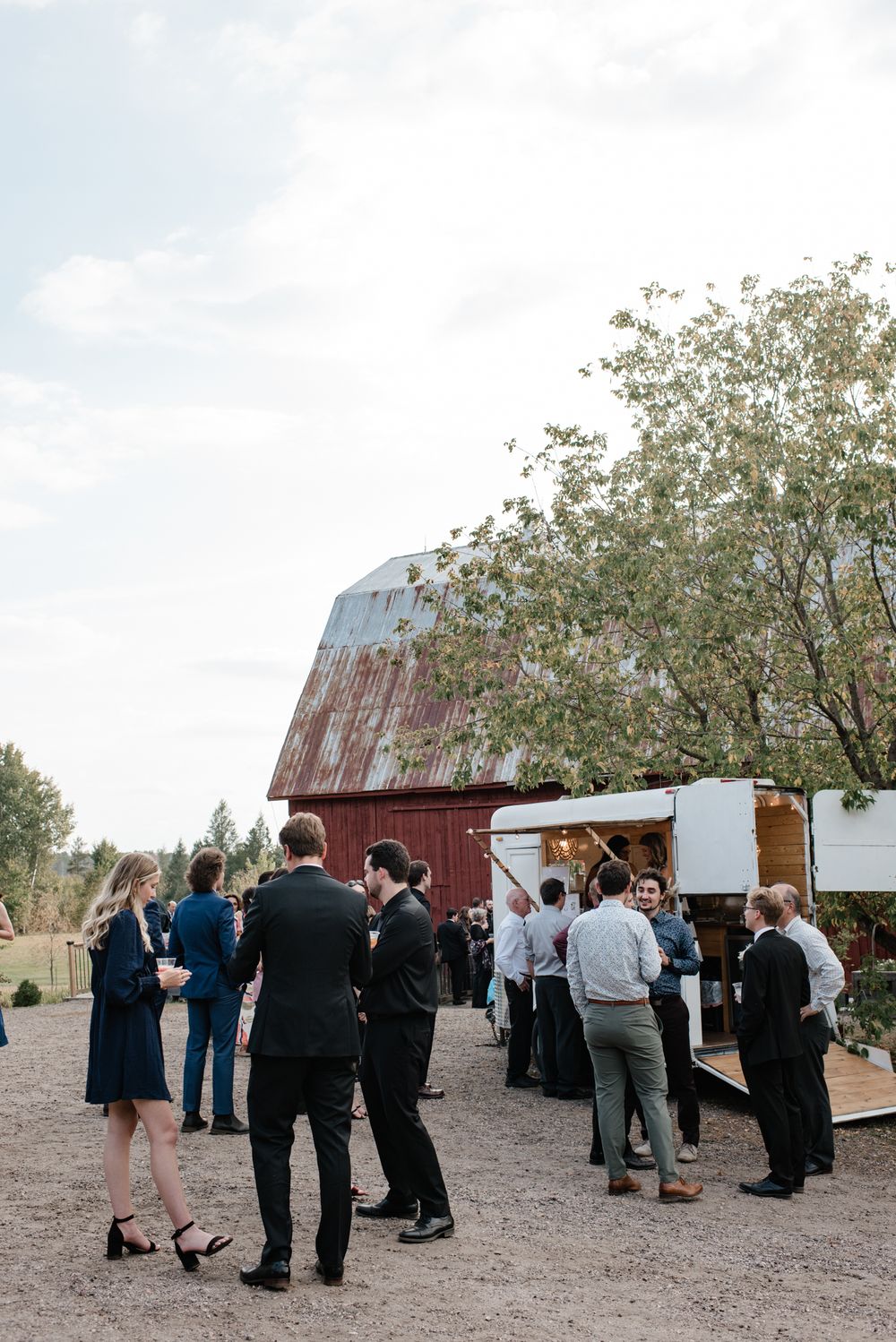 Guests mingle outdoors near a rustic red barn and vintage bar trailer at a countryside wedding reception.