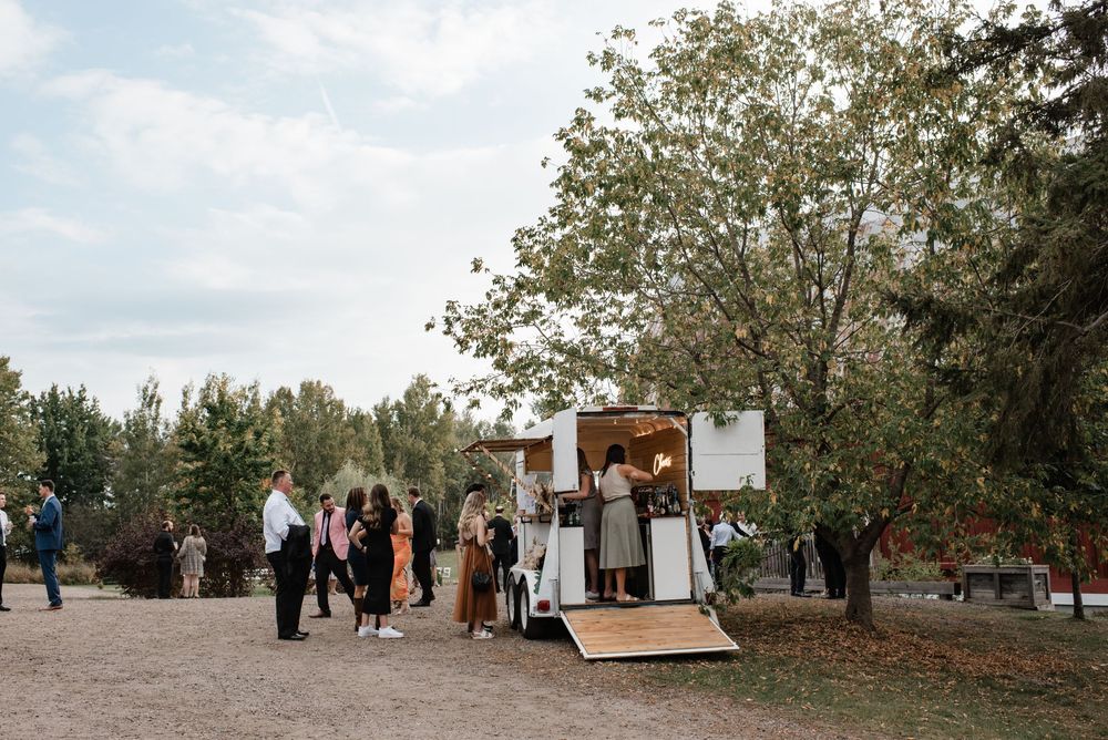 Wedding guests gather around a charming outdoor bar trailer beneath trees at an elegant rustic venue.