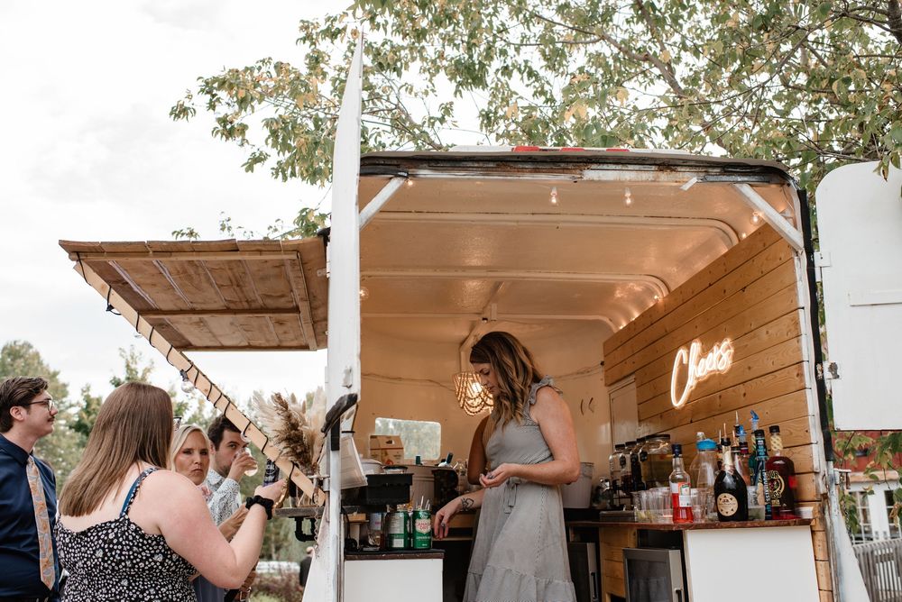 Guests enjoying drinks at a charming mobile bar with neon sign at an outdoor wedding reception.