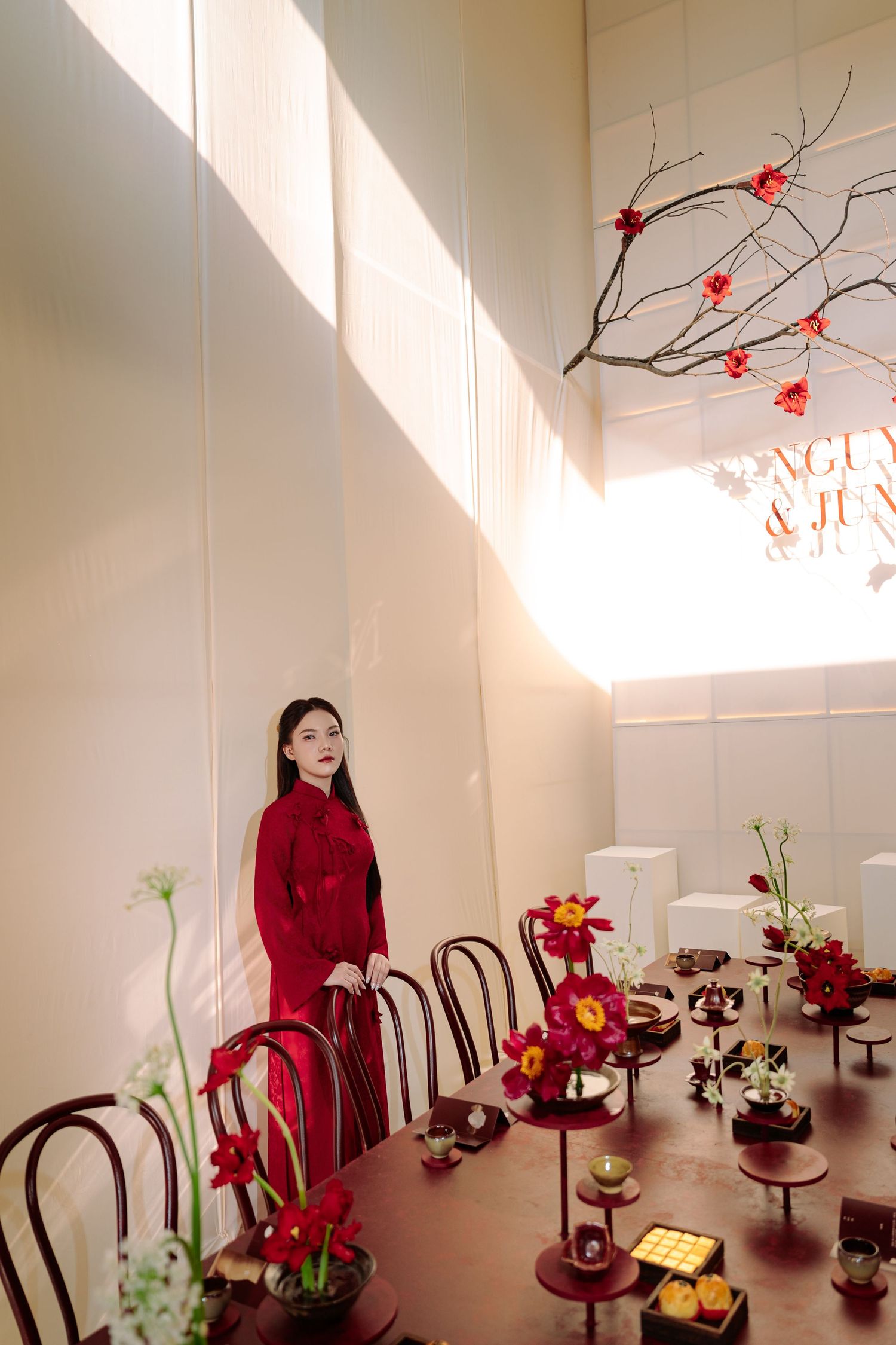 Bride in red ao dai standing beside decorated table with red flowers and traditional wedding items.