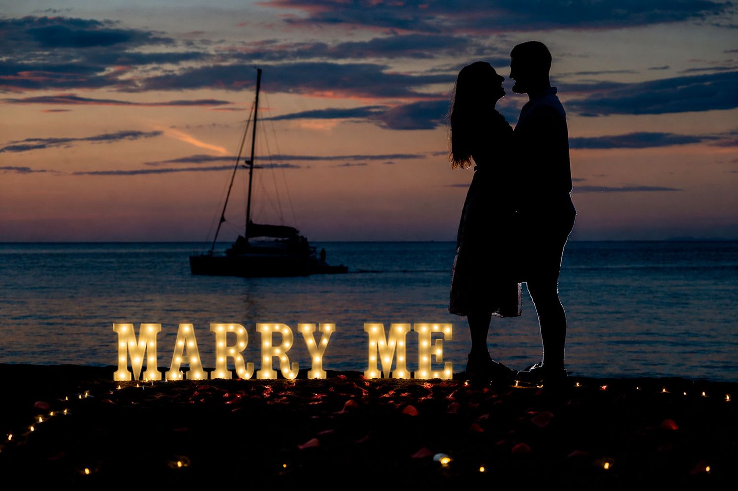 Romantic "Marry Me" letter sign on a beach at sunset with boats in the background.