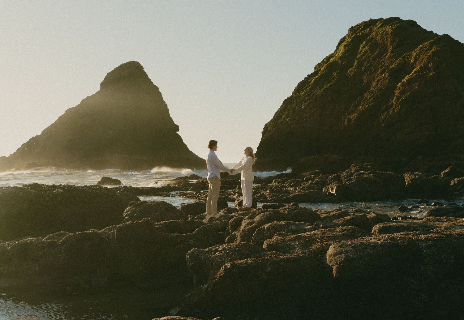 Couple holding hands on rocky Oregon Coast cliffs near Heceta Head Lighthouse during golden hour elopement session.