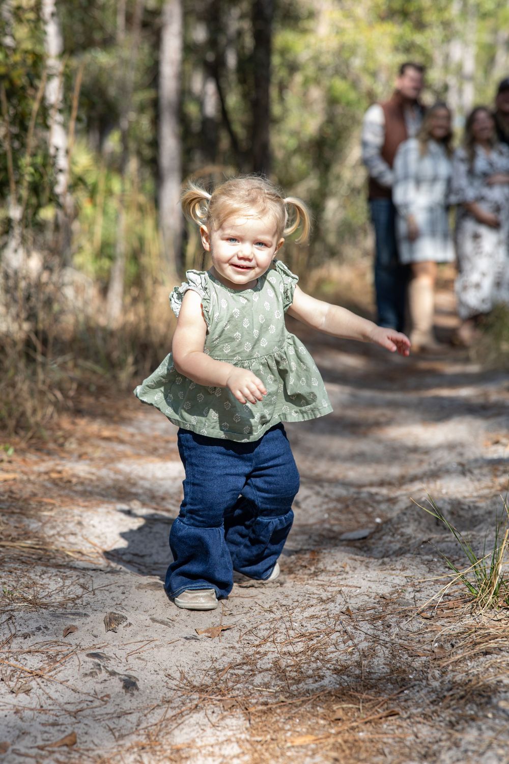 Toddler girl in green top and jeans running on a forest path with family in background.