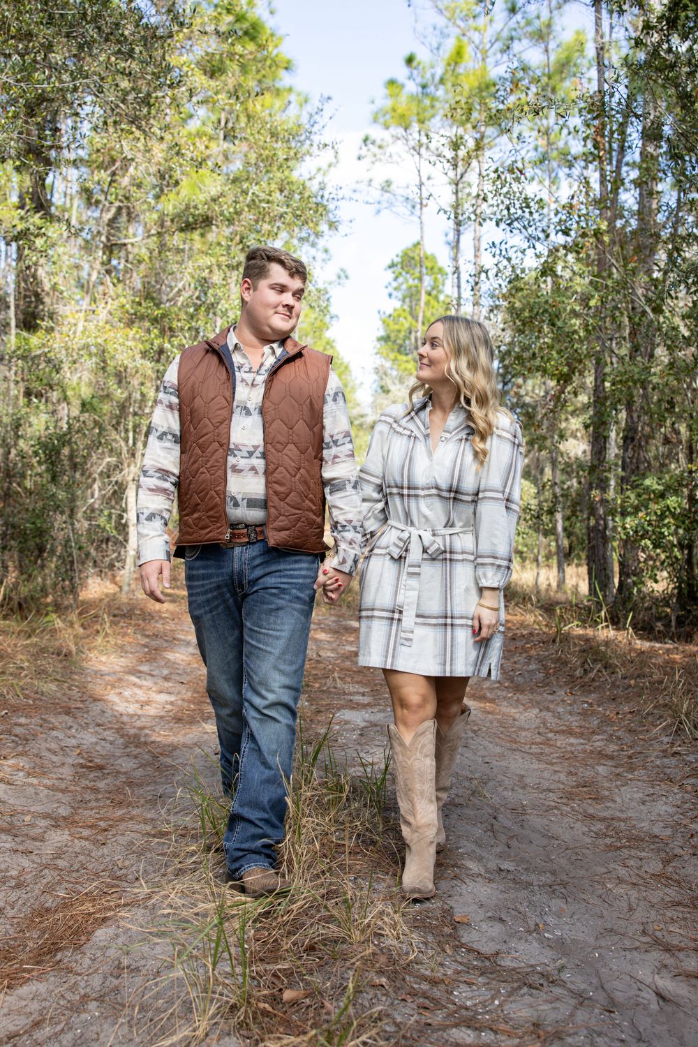 A couple walks hand in hand on a forest path, dressed in fall attire, smiling at each other.