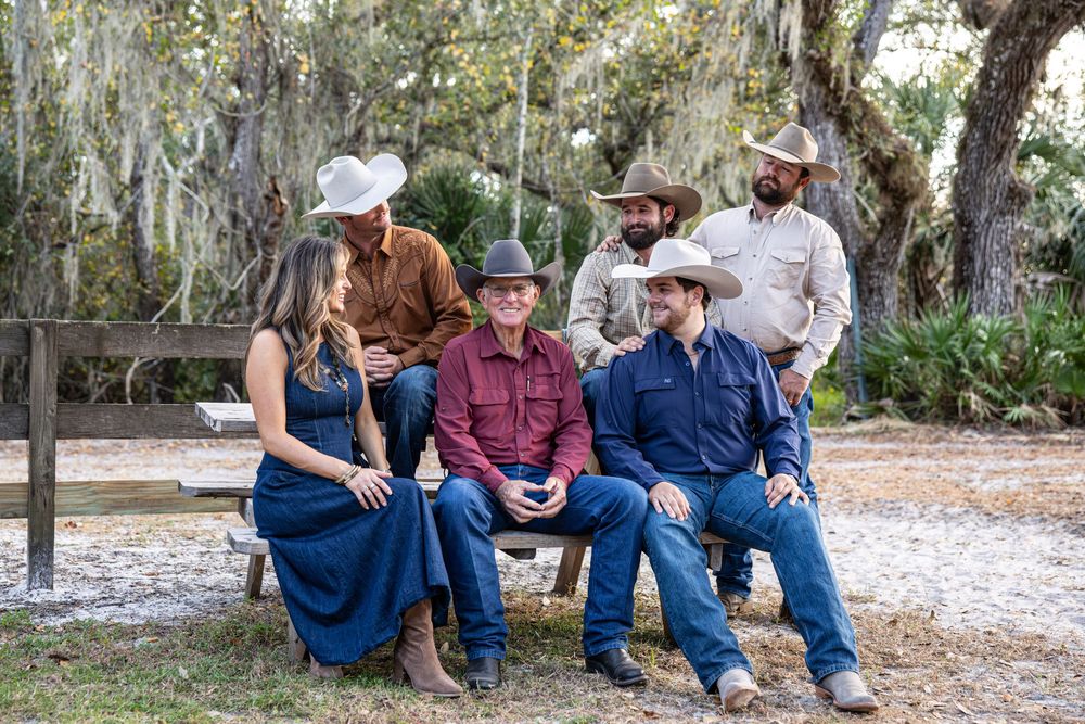 Group of five people in cowboy attire relaxing outdoors under Spanish moss-draped trees in a rustic setting.