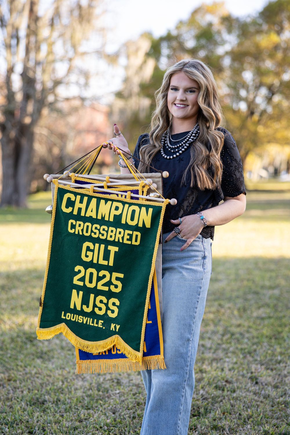 Young woman holding Champion Crossbred Gilt 2025 NJSS Louisville KY banner at outdoor livestock competition.