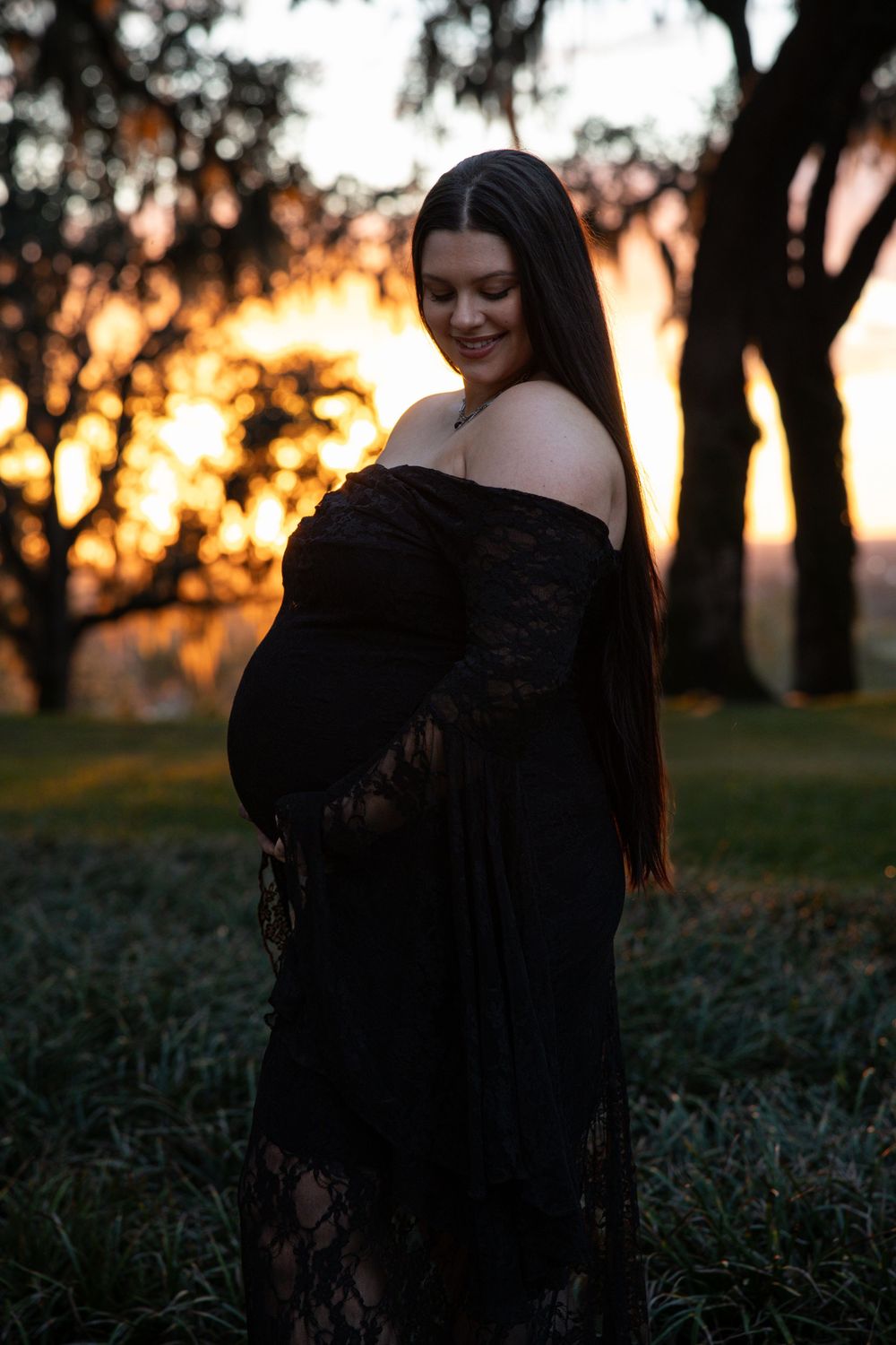 Pregnant woman in black lace off-shoulder gown poses outdoors at golden sunset, cradling her baby bump among trees.