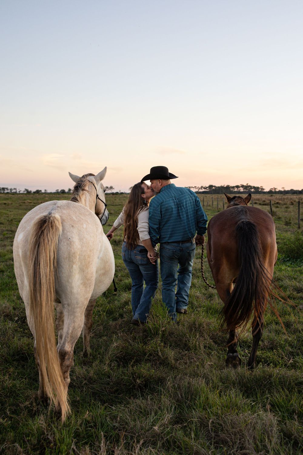 A couple shares a romantic moment at sunset in a field, standing between a white and brown horse.