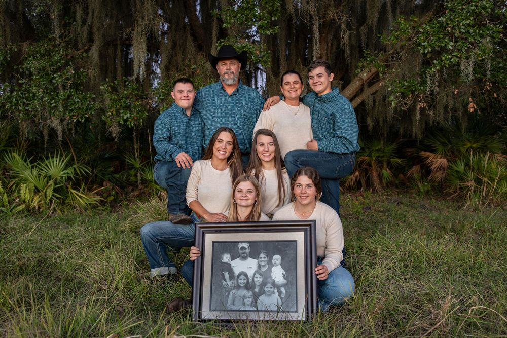 Family of seven poses outdoors under Spanish moss trees, holding a vintage black-and-white family portrait photo.