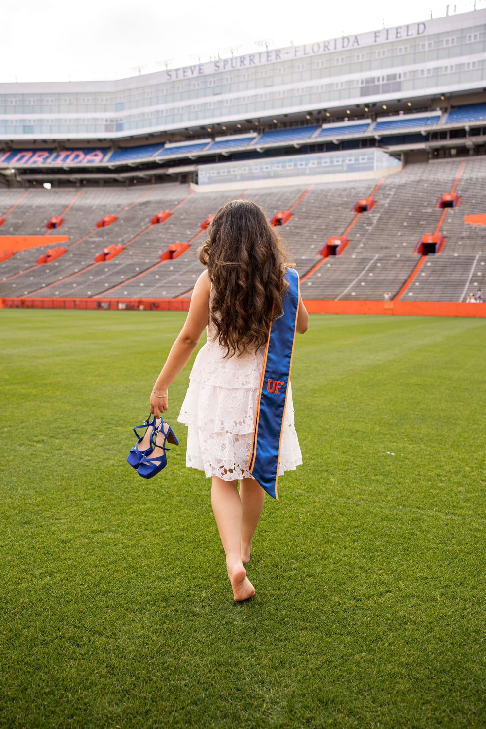 A University of Florida graduate walks barefoot on the stadium field, holding blue heels and wearing a UF graduation stole.
