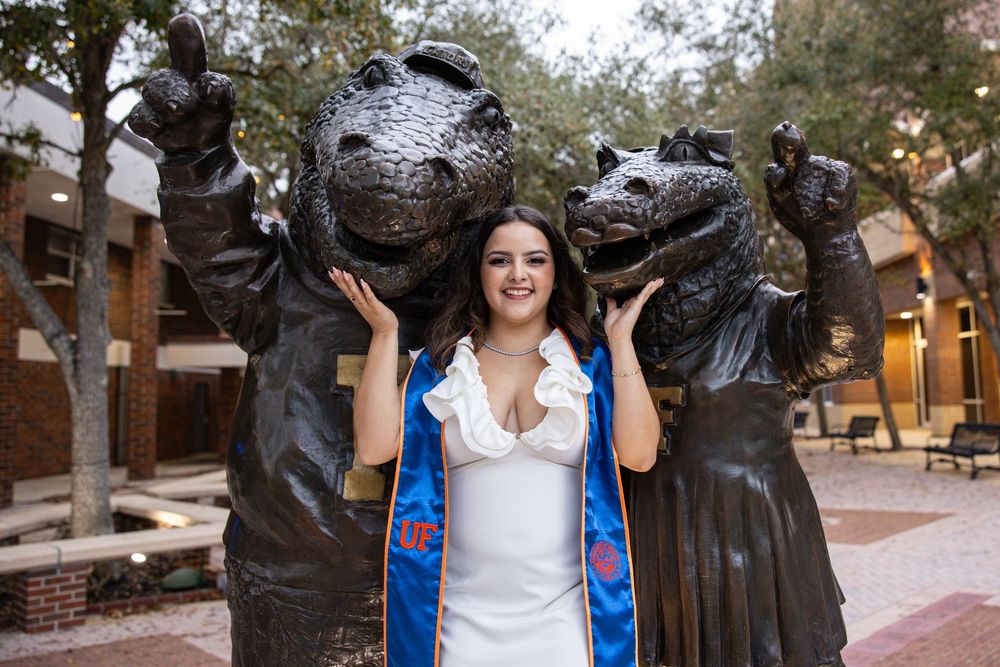 Graduate in white dress and blue stole poses between two bronze hippo statues at University of Florida campus.