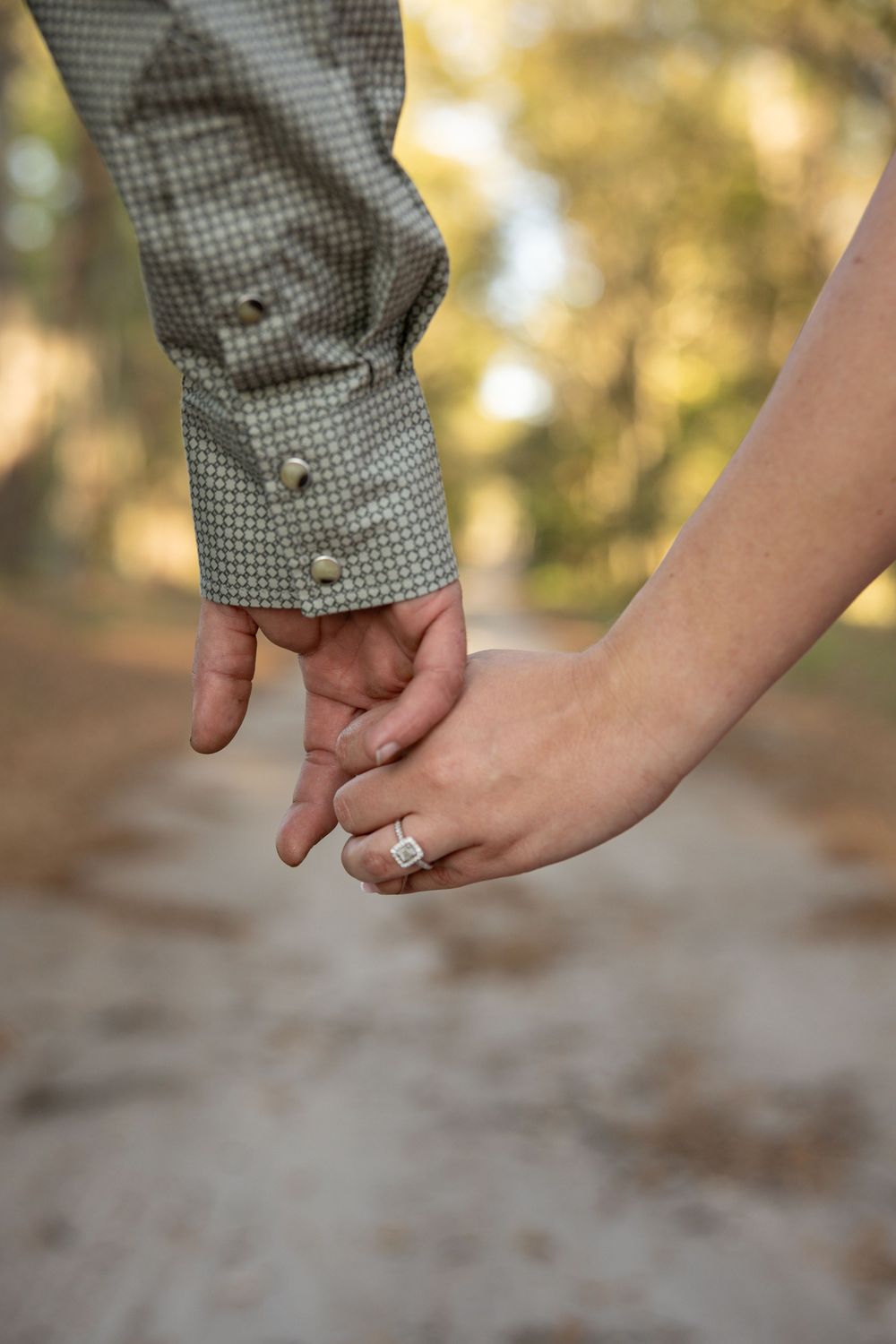 Couple holding hands on a forest path, woman wearing a diamond engagement ring, romantic outdoor setting.