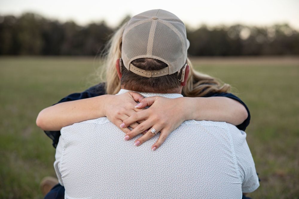 A woman with blonde hair hugs a man in a tan cap from behind in an outdoor field setting.