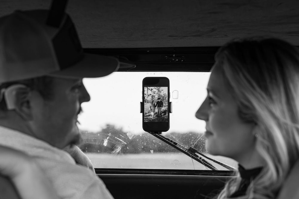 Black and white photo of two people in a car with a phone mounted on the windshield displaying a photo.