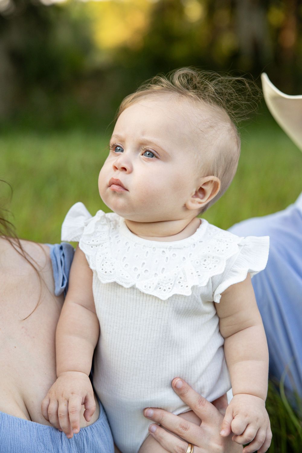 Adorable baby girl in white ruffle onesie with blue eyes sitting outdoors in green park setting.
