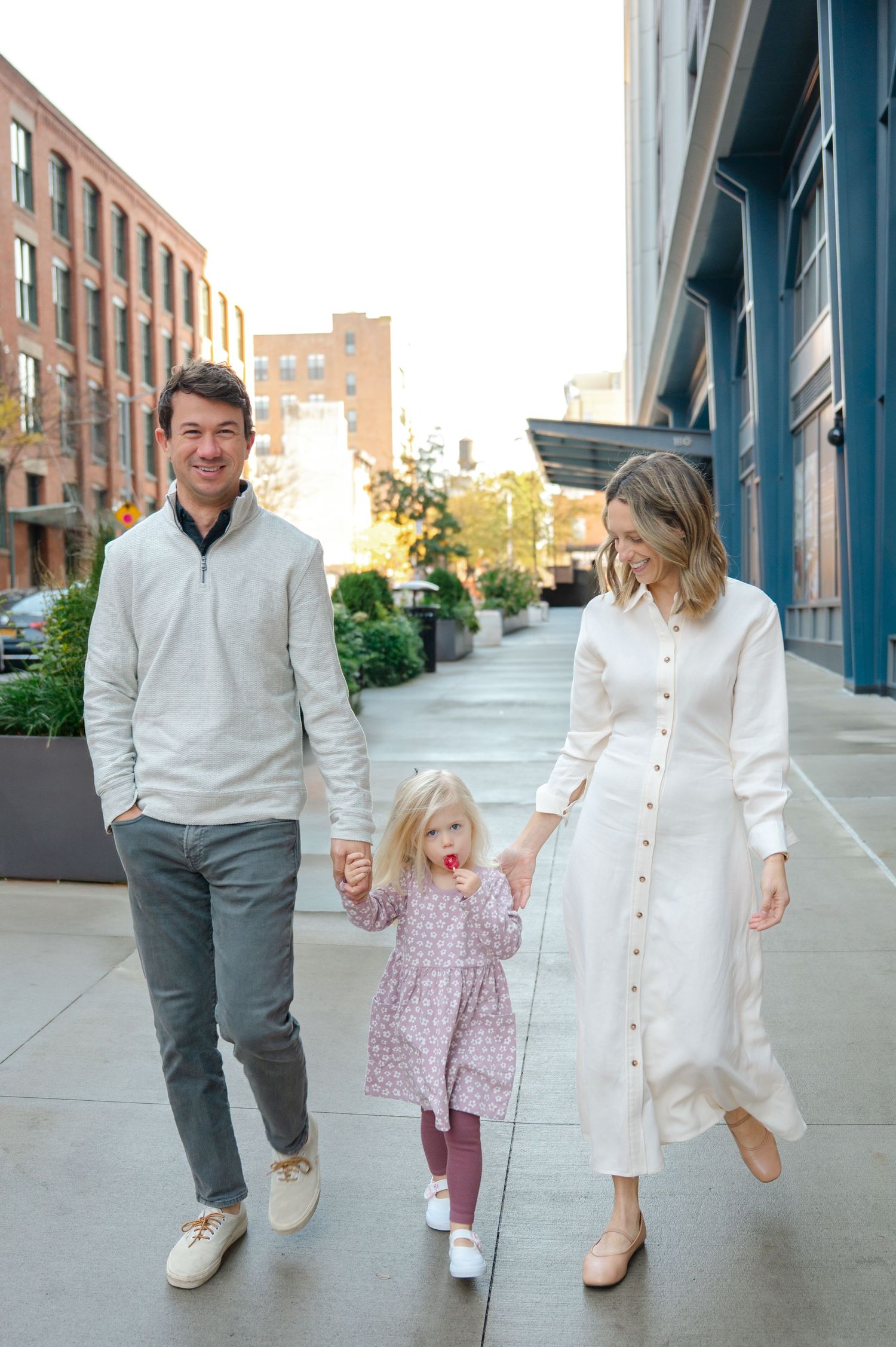 A happy family of three walks hand-in-hand on a city sidewalk, parents in white outfits with a young blonde girl in a pink dress.