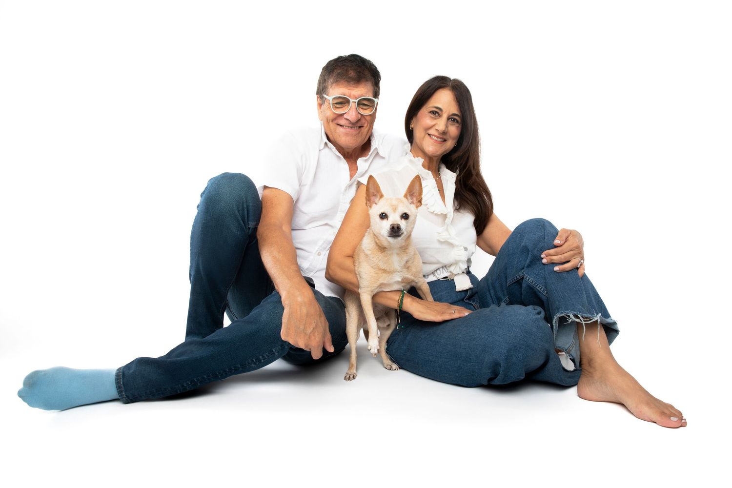 A smiling couple sitting on white background with a small tan Chihuahua dog between them, wearing casual jeans and white shirts.