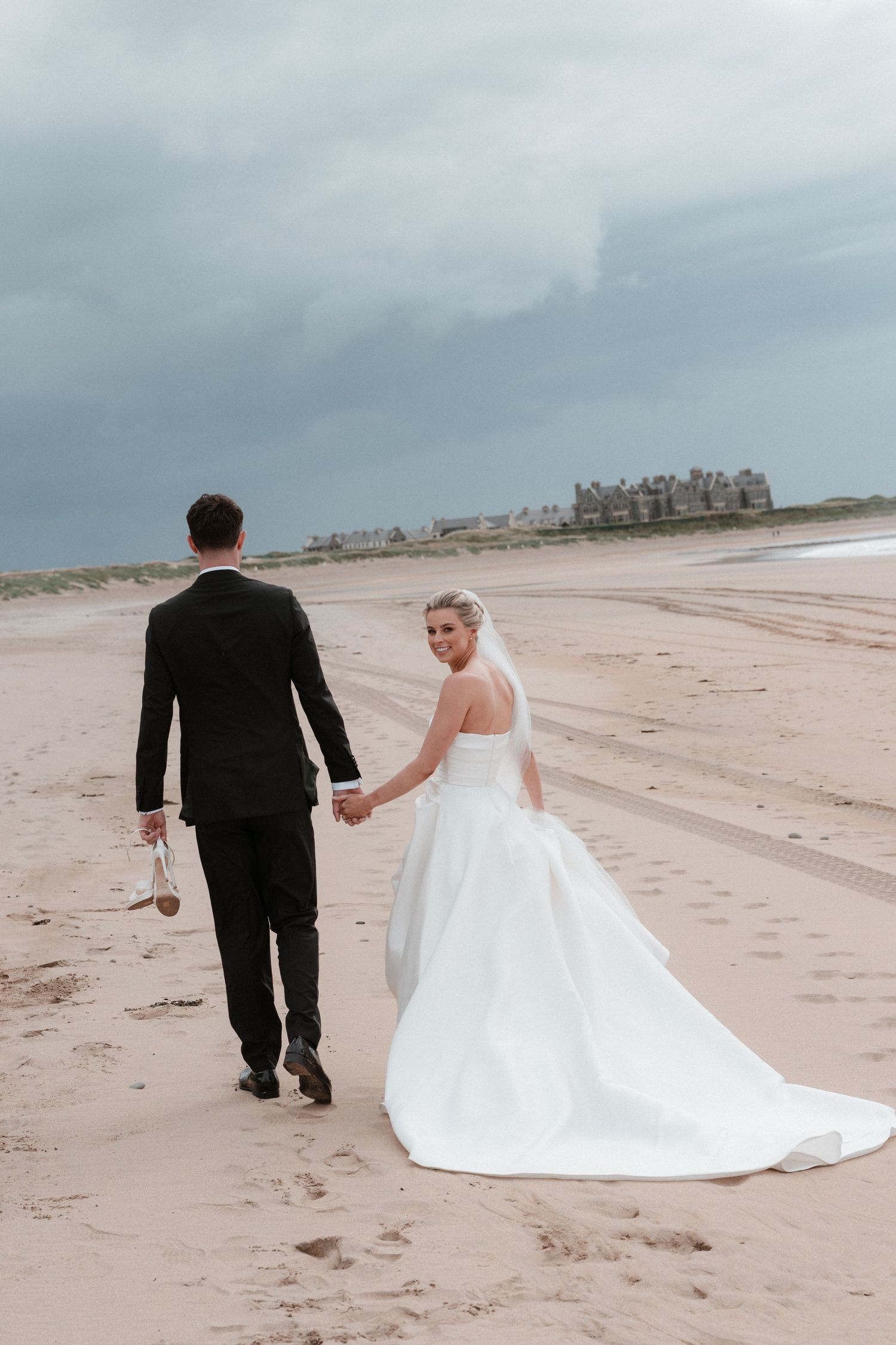 Bride in white gown looks back smiling while holding groom's hand walking on sandy beach under cloudy skies.
