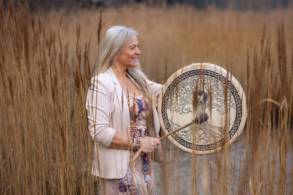 Woman holding decorative drum with tree design standing in golden reed field wearing floral dress and white blazer.