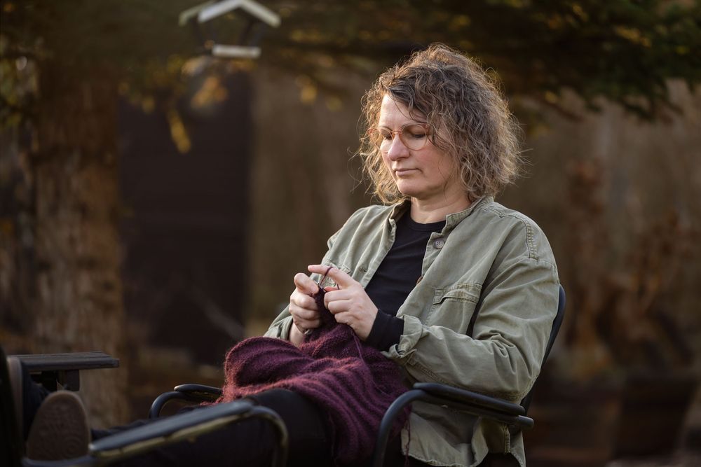 Woman with curly hair knitting a burgundy project on an outdoor bench surrounded by autumn foliage.