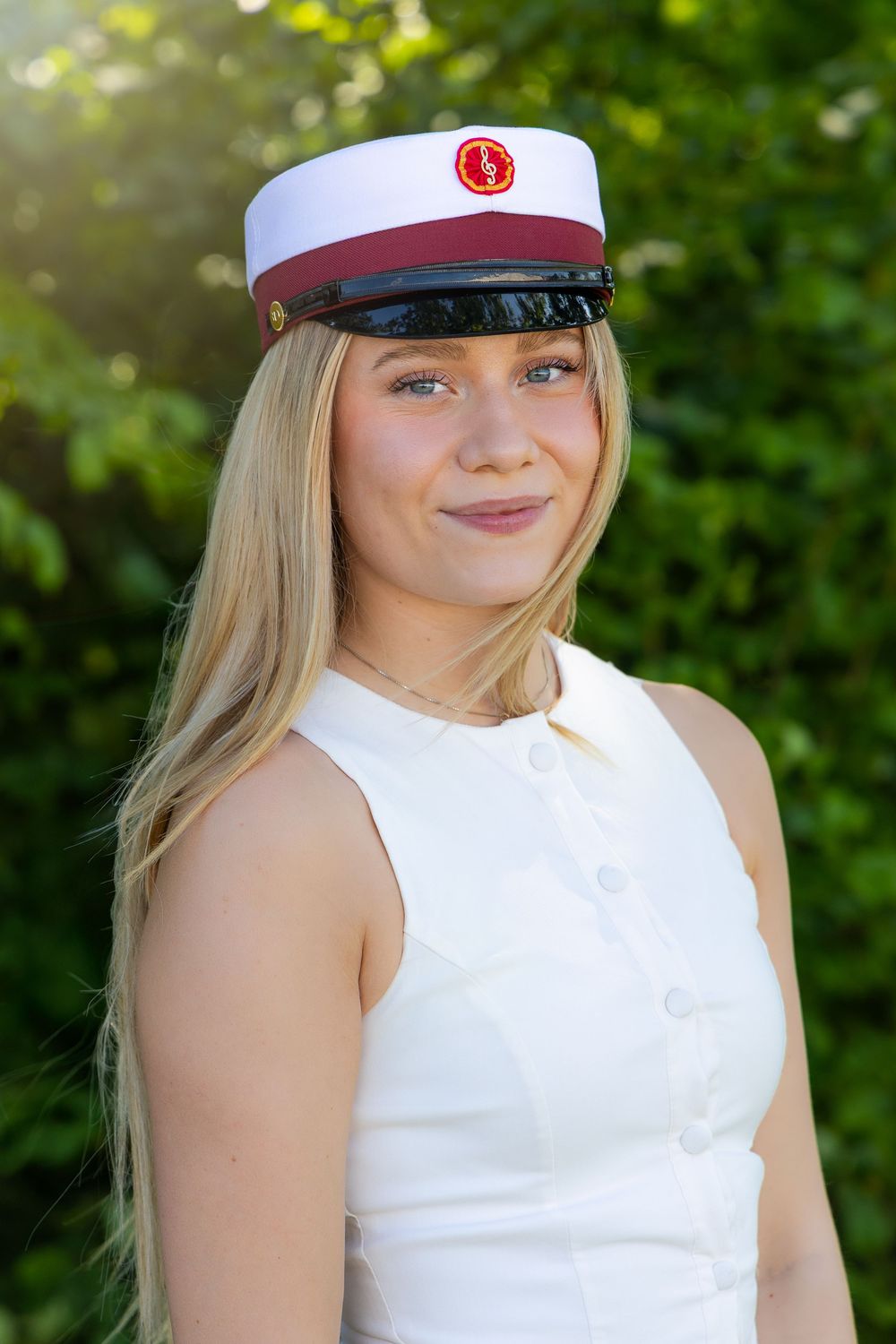 Young blonde woman smiling in white dress and burgundy graduation cap outdoors with green trees background.