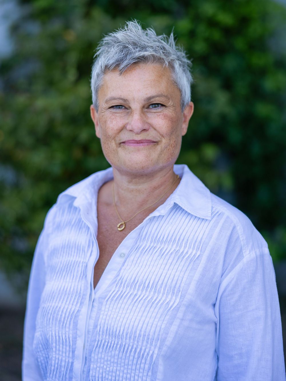A smiling middle-aged woman with short gray hair wearing a white linen shirt, standing outdoors with green foliage background.