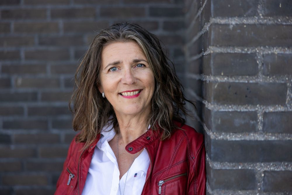 Smiling middle-aged woman with brown hair wearing a red leather jacket, posing outdoors near a brick wall.