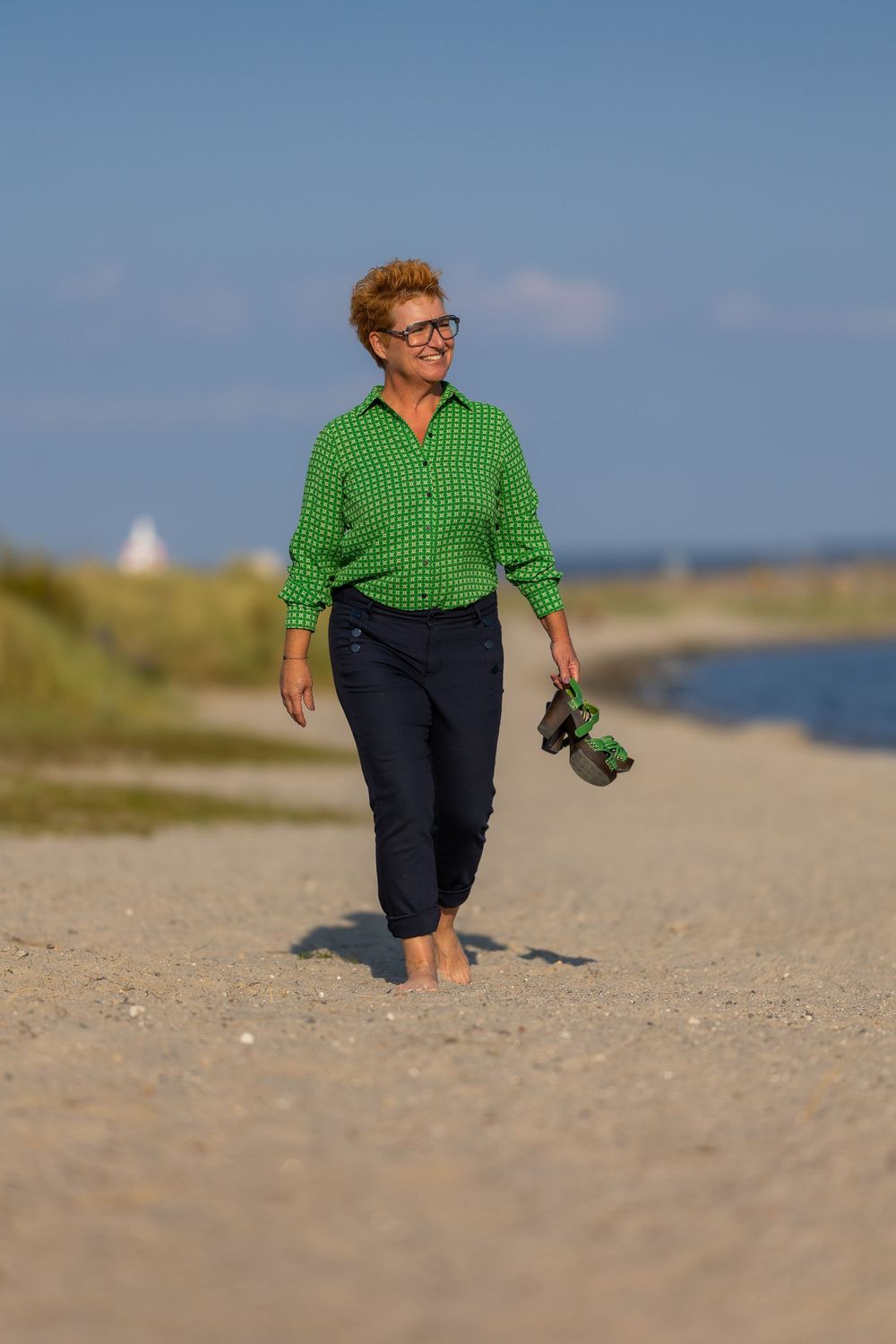 Middle-aged woman in green shirt walks barefoot on sandy beach carrying shoes on a sunny day.