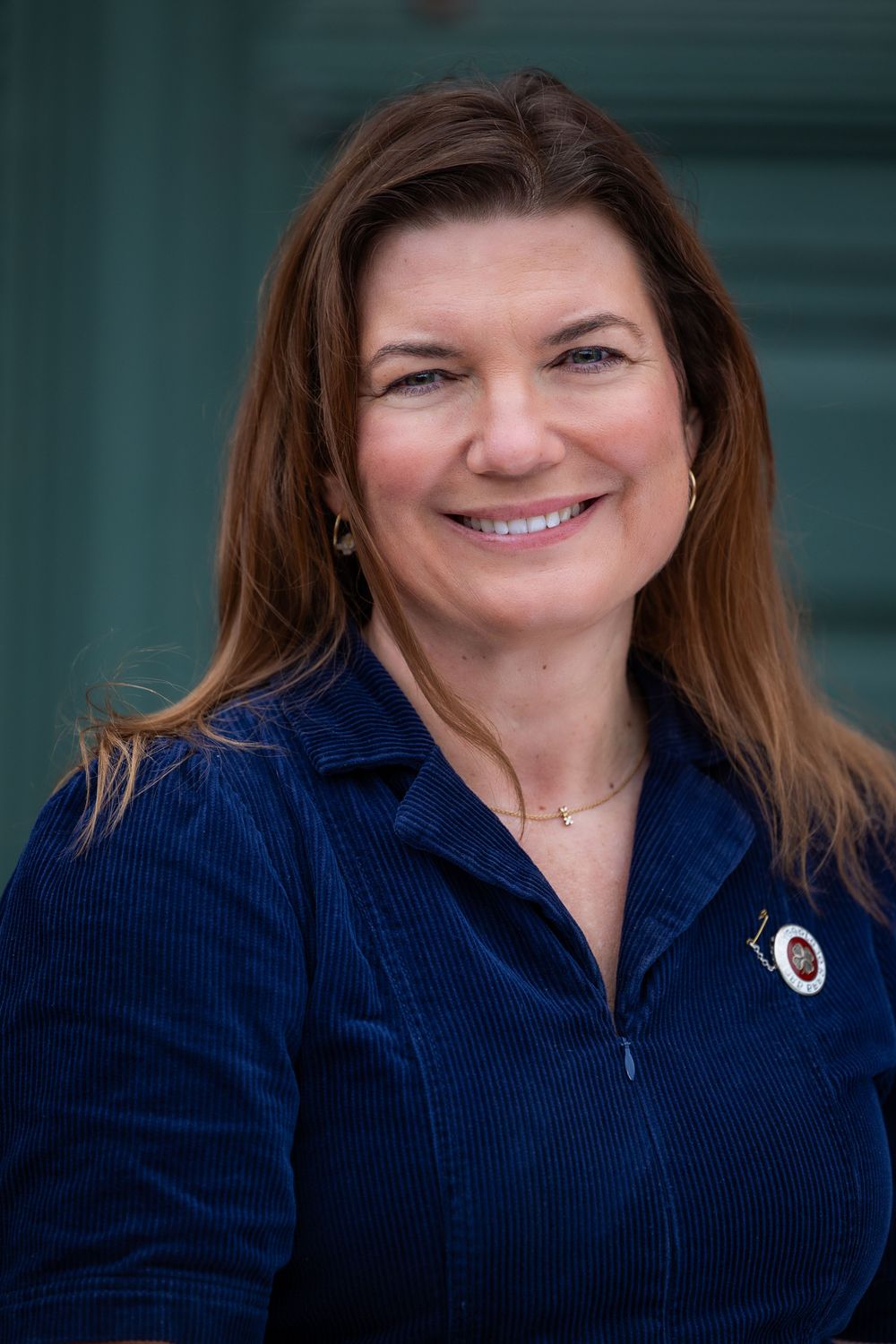 Smiling woman with brown hair wearing a blue corduroy jacket and small pin, posed in front of a teal door.