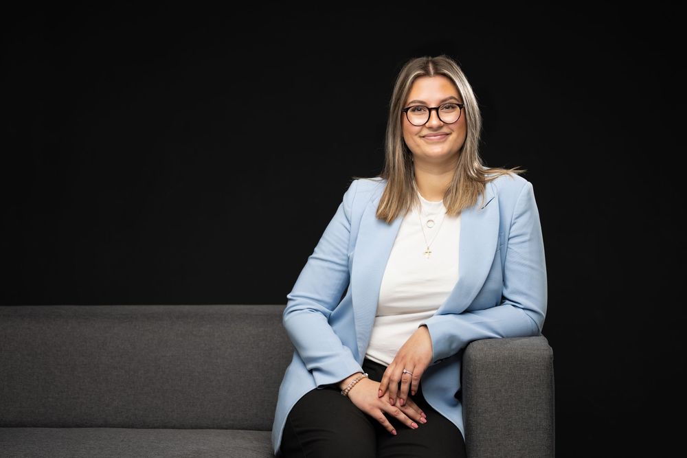 Professional woman in light blue blazer and glasses sitting on gray sofa against dark background.