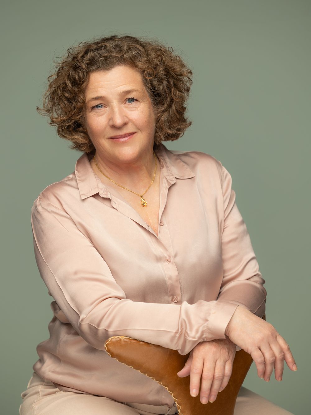 A middle-aged woman with curly hair wearing a blush pink blouse sits relaxed on a stool against a sage green background.