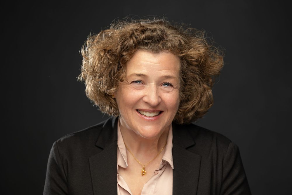 Professional headshot of a smiling middle-aged woman with curly hair wearing a black blazer against a dark background.