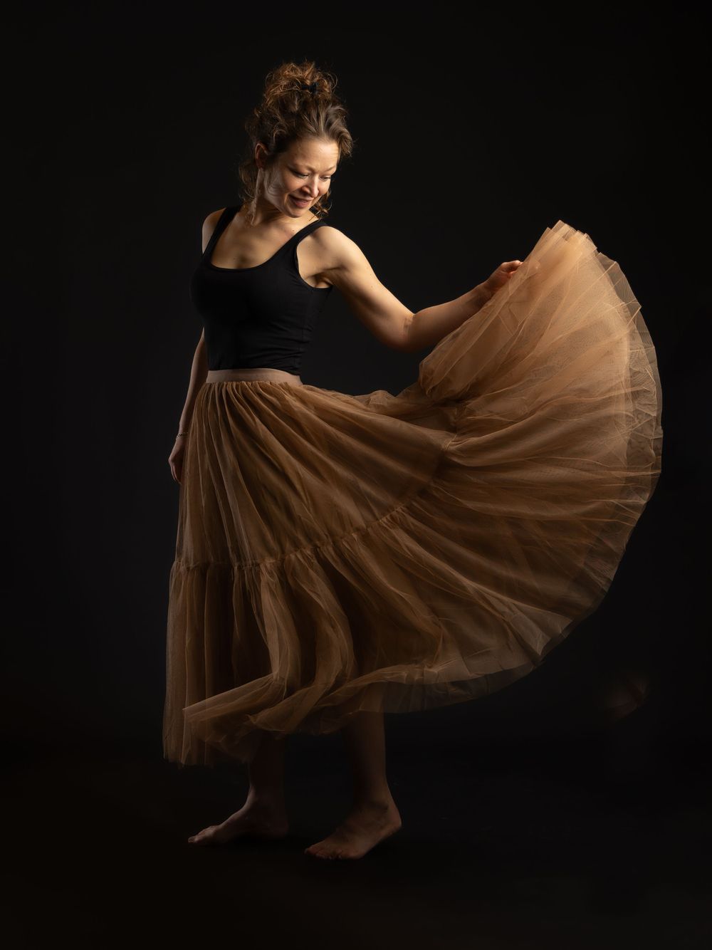 A woman twirls in a flowing golden tulle skirt and black tank top, smiling against a dark studio background.