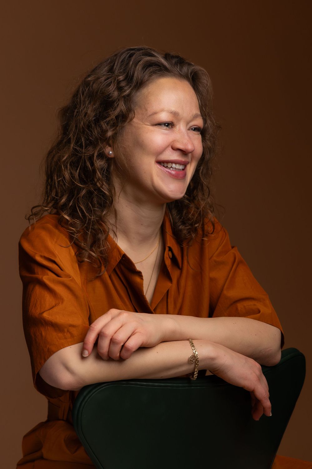 Smiling woman with curly hair wearing an orange shirt, seated on a green chair against a warm brown background.