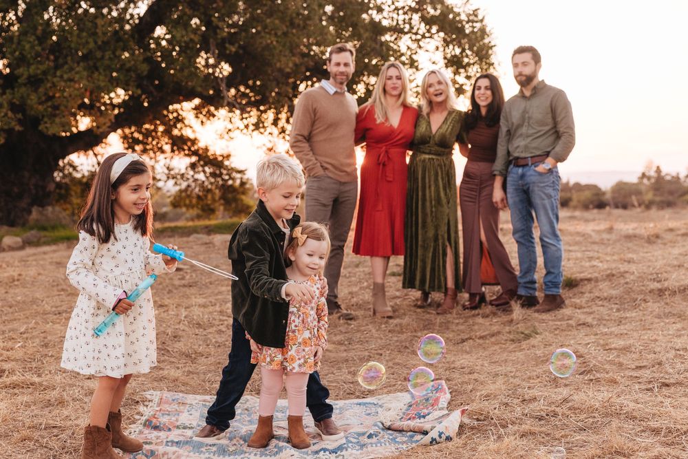 Family portrait at golden hour outdoors, children playing with bubbles while adults pose near a tree.