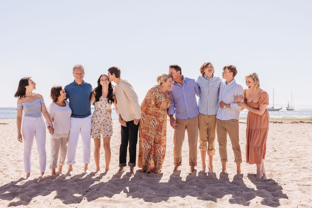 Large family group posing together on sunny beach, smiling and embracing in pastel outfits with ocean in background.