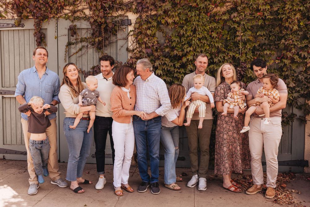 Multi-generational family portrait outdoors, adults and young children posing together against ivy-covered wall in warm neutral tones.