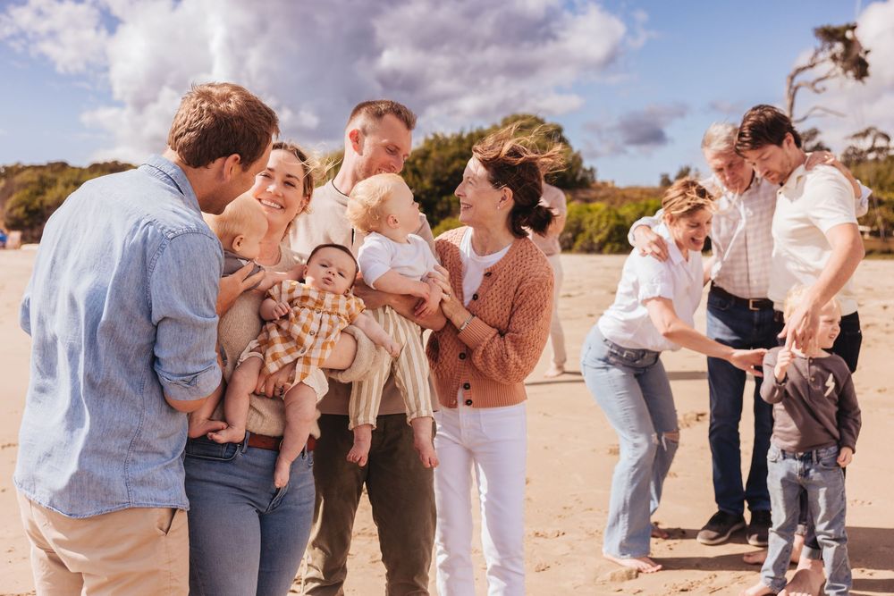 Large family gathering on a sandy beach, sharing joyful moments with babies and toddlers on a sunny day.