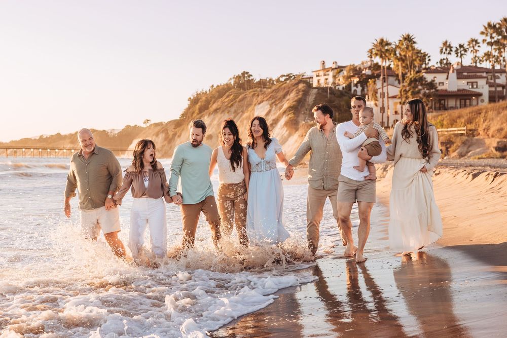Family group walking along sunny beach shoreline at golden hour, waves splashing, wearing coordinated white and neutral outfits.