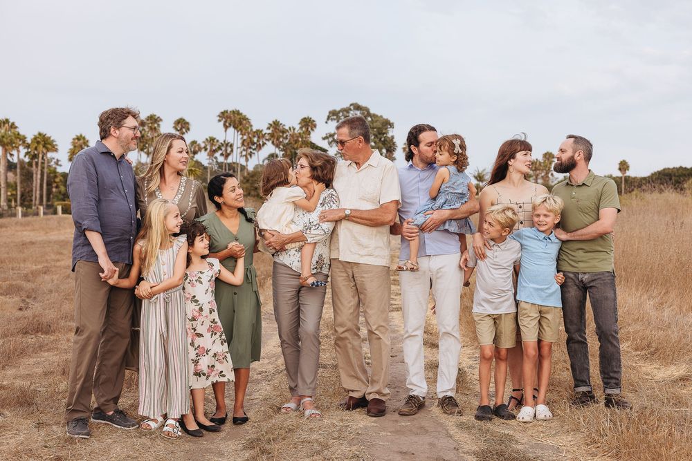 Large extended family posing together outdoors in a golden field with palm trees, dressed in casual earth-toned outfits.