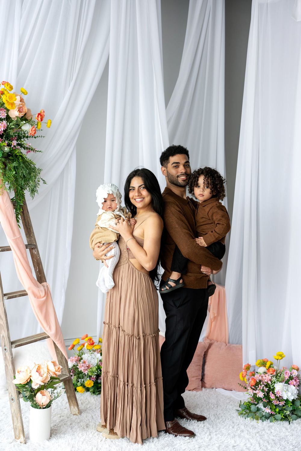 A joyful family of four poses in a floral studio setting, parents holding two young children amid white drapes and colorful flowers.