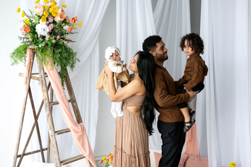 A family of four shares a candid moment near a floral ladder decoration with white curtain backdrop in a photography studio.