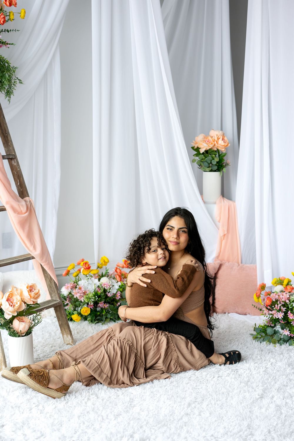 A mother and her curly-haired toddler share a tender embrace on a white rug surrounded by colorful flowers in a studio.