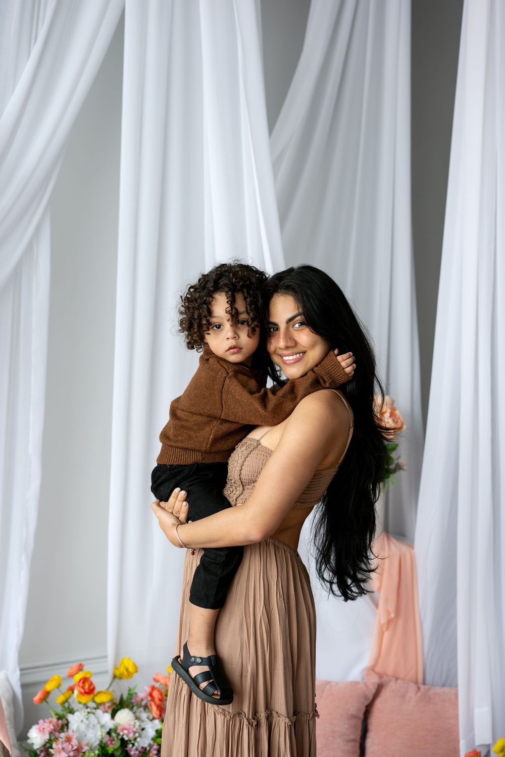 A smiling mother in a tan dress holds her curly-haired toddler in a brown jacket against white drapes.