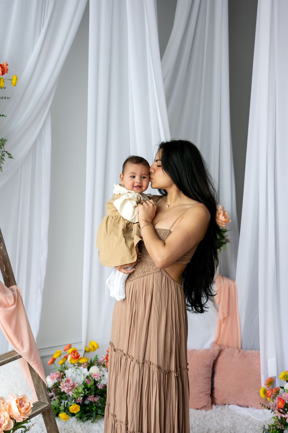 A dark-haired mother in a tan dress kisses her infant baby in a gold dress near colorful flowers.