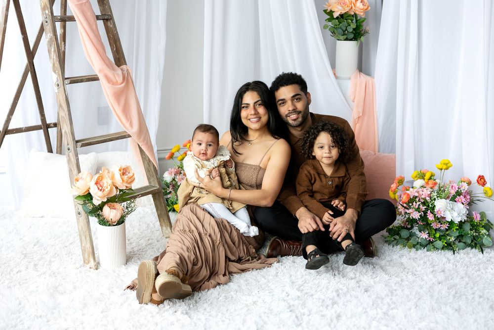 A happy family of four poses together on a white fluffy rug surrounded by colorful floral arrangements.