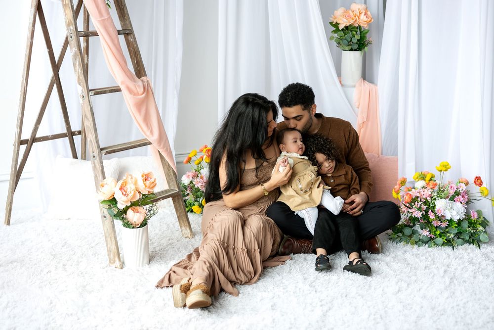 A loving family sits together on a white rug as parents kiss their baby while their toddler watches.