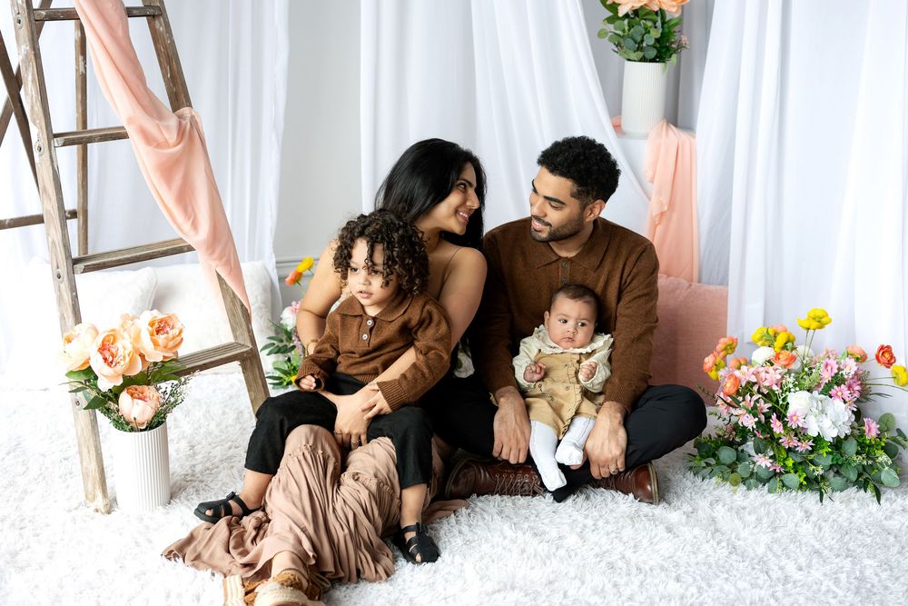 A couple with two young children share a tender moment sitting on a white rug with floral decor.