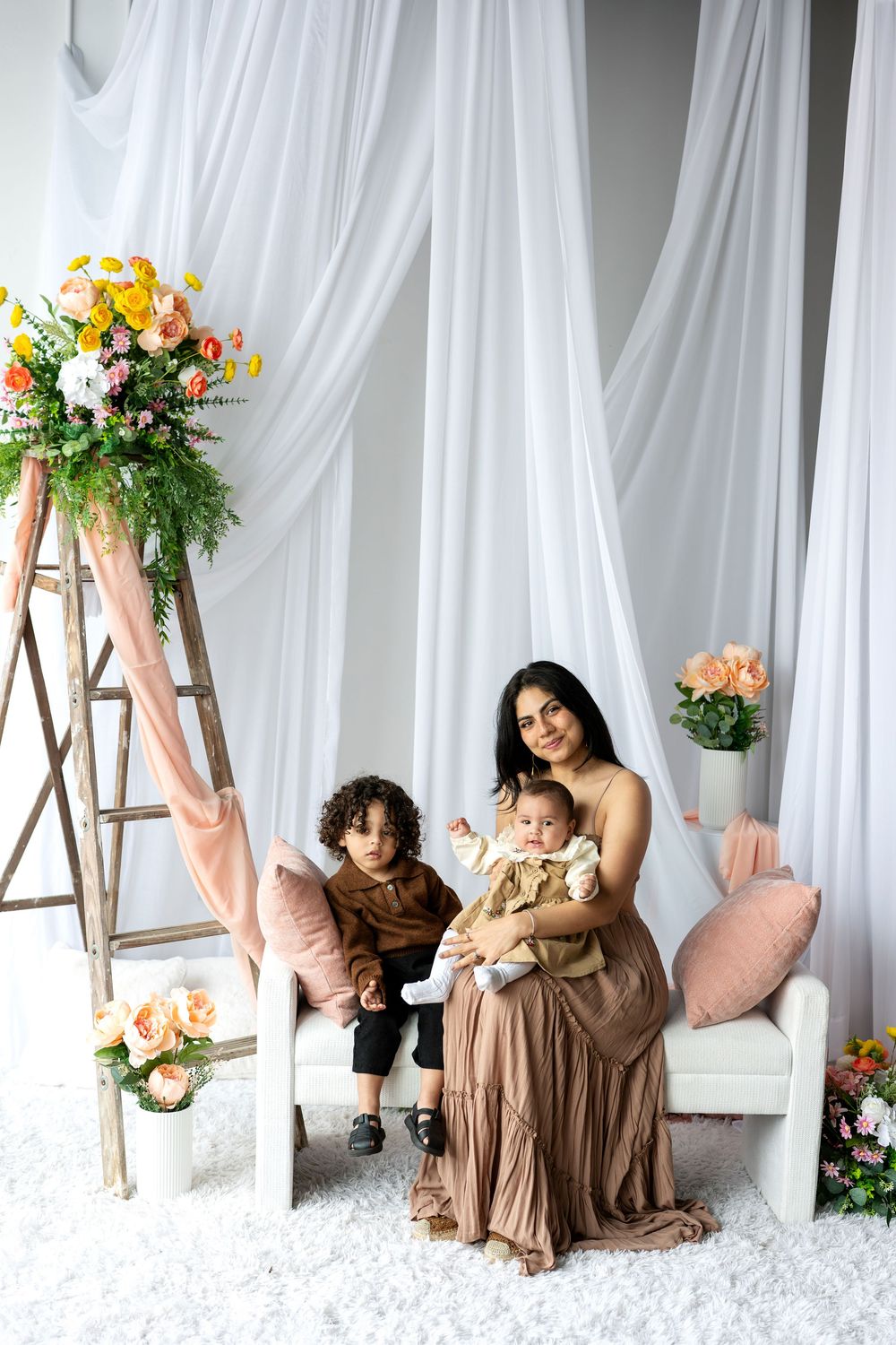 A mother in a brown dress poses with her toddler and baby in a floral studio with white drapes.