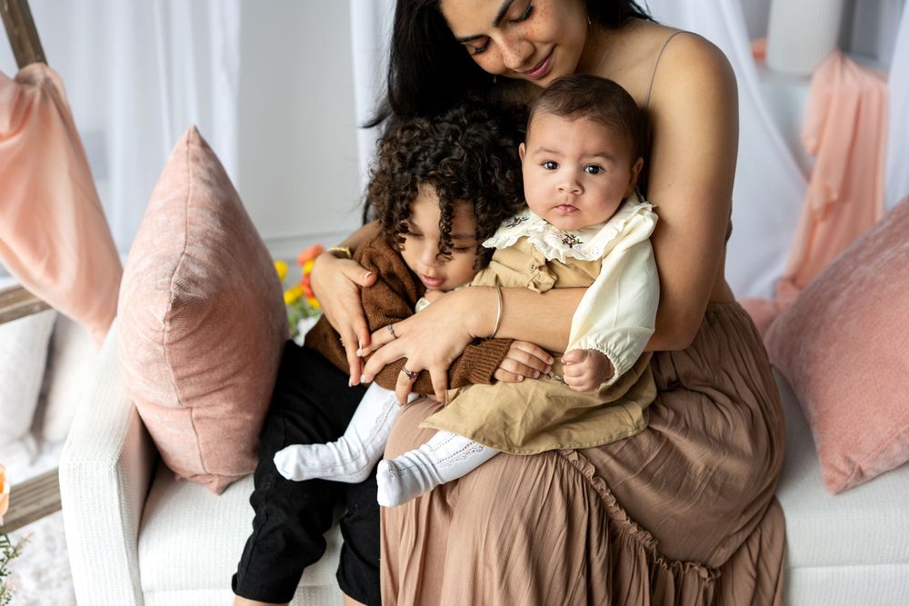 A mother in a tan dress cuddles her toddler and infant baby on a white bench with pink pillows.